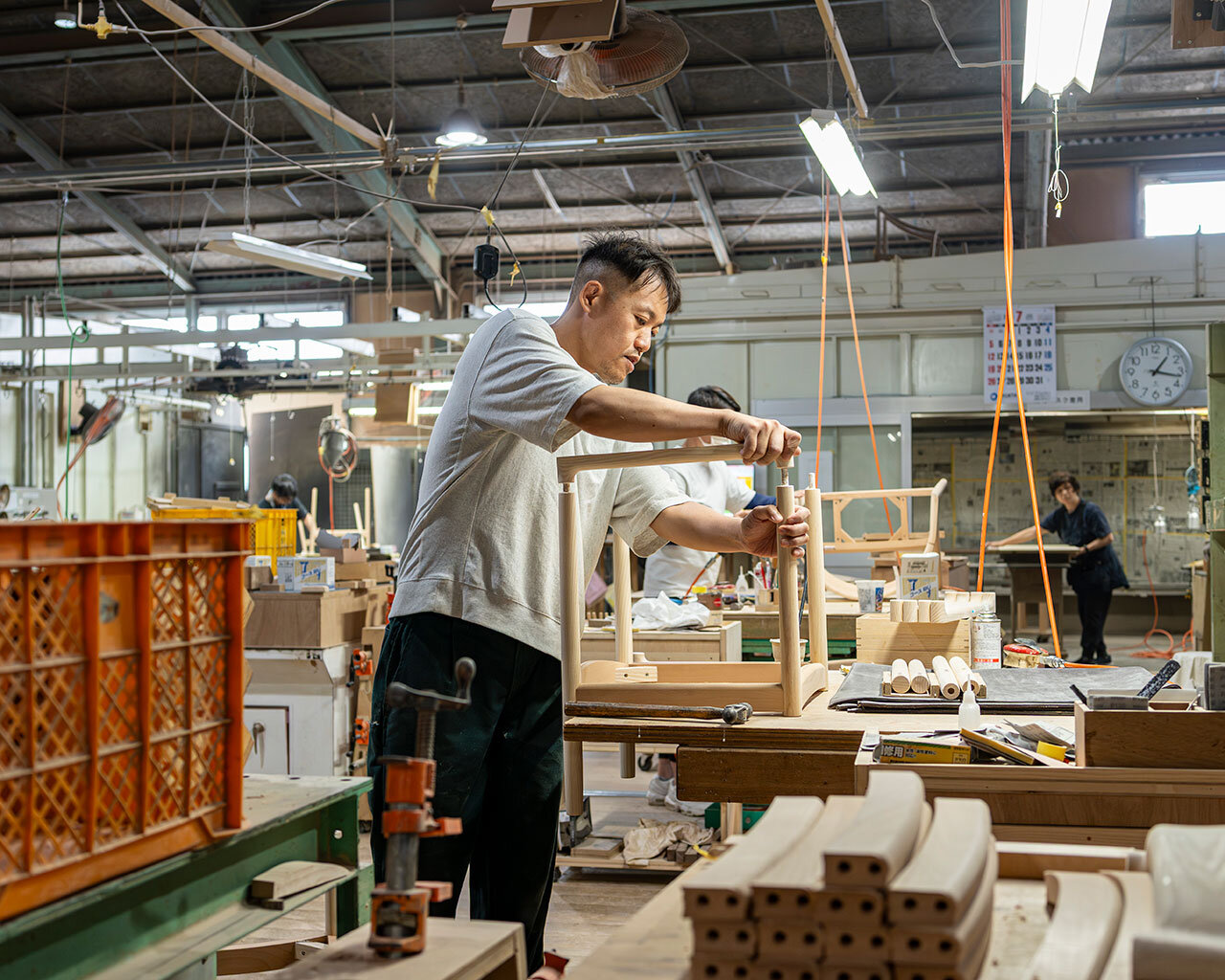 徳島の家具工場で、組み上がった木製椅子の接合部や仕上がりを入念にチェックする若手家具職人。丁寧な品質管理と無垢家具づくりのリアルな現場風景。